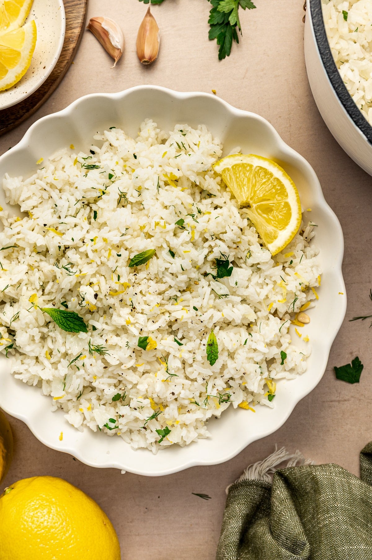 Greek lemon rice served in a white scalloped bowl with lemon slices, lemon zest, chopped parsley, dill, mint leaves, and black pepper. A cream-colored Dutch oven, garlic cloves, parsley, lemon wedges, whole lemons, and a green napkin are visible around the bowl.