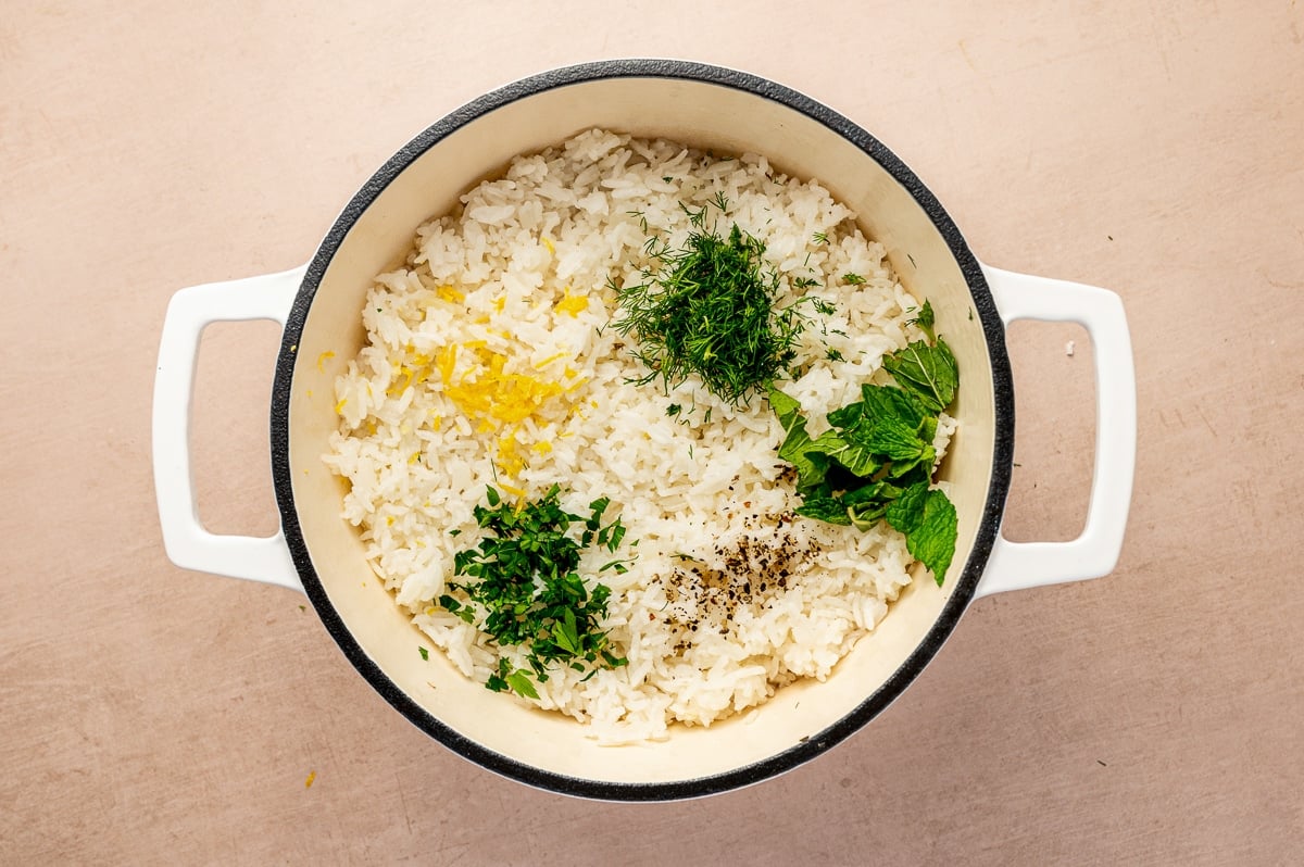 Cooked white rice in a cream-colored Dutch oven topped with lemon zest, chopped parsley, fresh dill, mint leaves, and black pepper before being mixed together.