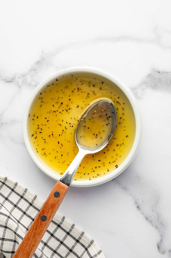 Golden honey-lime dressing with black pepper in a small white bowl, with a metal spoon with a wooden handle resting inside on a white marble countertop.