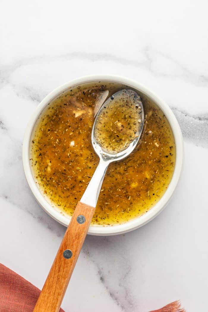 Overhead close-up of lemon oregano dressing in a small white bowl with a wooden-handled spoon, showing olive oil, minced garlic, dried oregano, lemon juice, red wine vinegar, salt, and black pepper on a white marble surface.