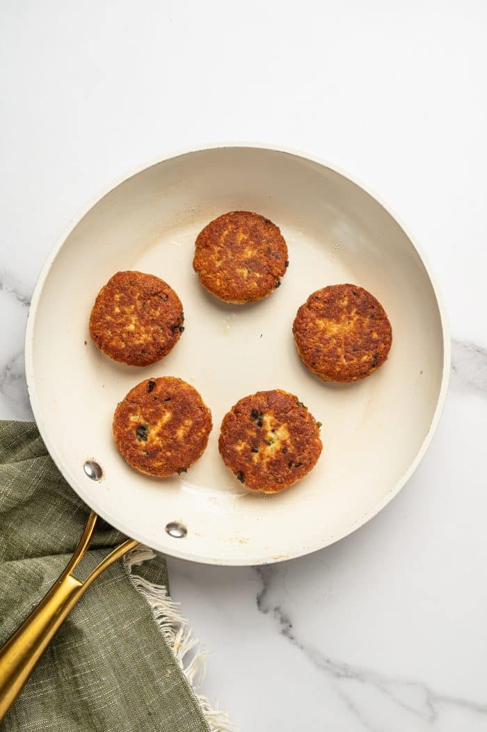 Five golden-brown salmon cakes are cooking in a cream-colored skillet with gold handles on a white marble surface, with an olive-green napkin beside the pan. The salmon cakes have crisp browned edges and visible green onion pieces.