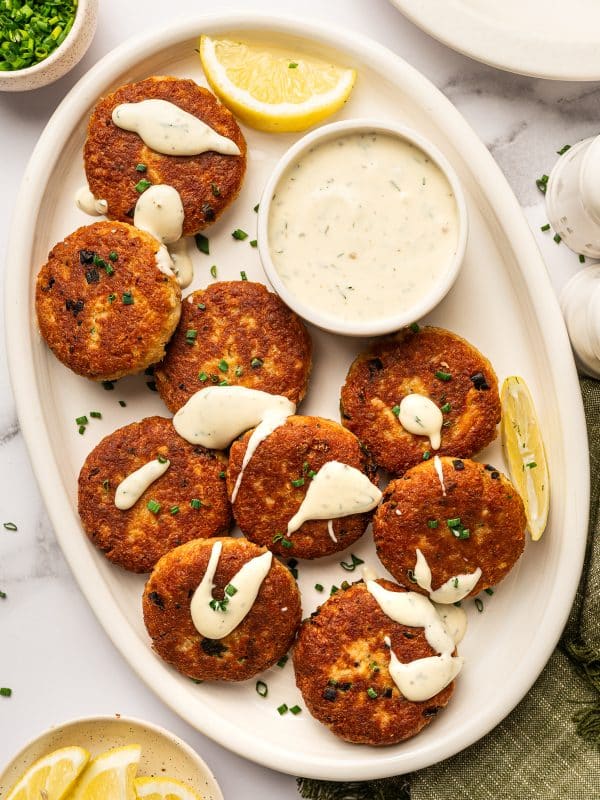 Overhead view of golden-brown salmon cakes on a cream oval platter with creamy white sauce drizzled over the tops, a small white bowl of sauce in the center, lemon wedges, chopped green chives, an olive-green linen napkin, and a small bowl of lemon wedges on a white marble surface.