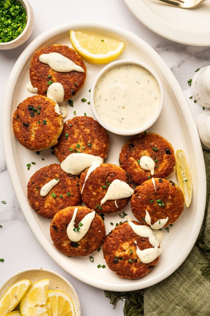 Overhead view of golden-brown salmon cakes on a cream oval platter with creamy white sauce drizzled over the tops, a small white bowl of sauce in the center, lemon wedges, chopped green chives, an olive-green linen napkin, and a small bowl of lemon wedges on a white marble surface.