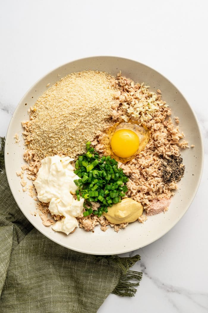 Ingredients for salmon cakes arranged in a beige-speckled bowl, including flaked canned salmon, plain breadcrumbs, mayonnaise, Dijon mustard, one egg, chopped green onions, minced garlic, salt, and black pepper, on a white marble surface with an olive-green napkin.