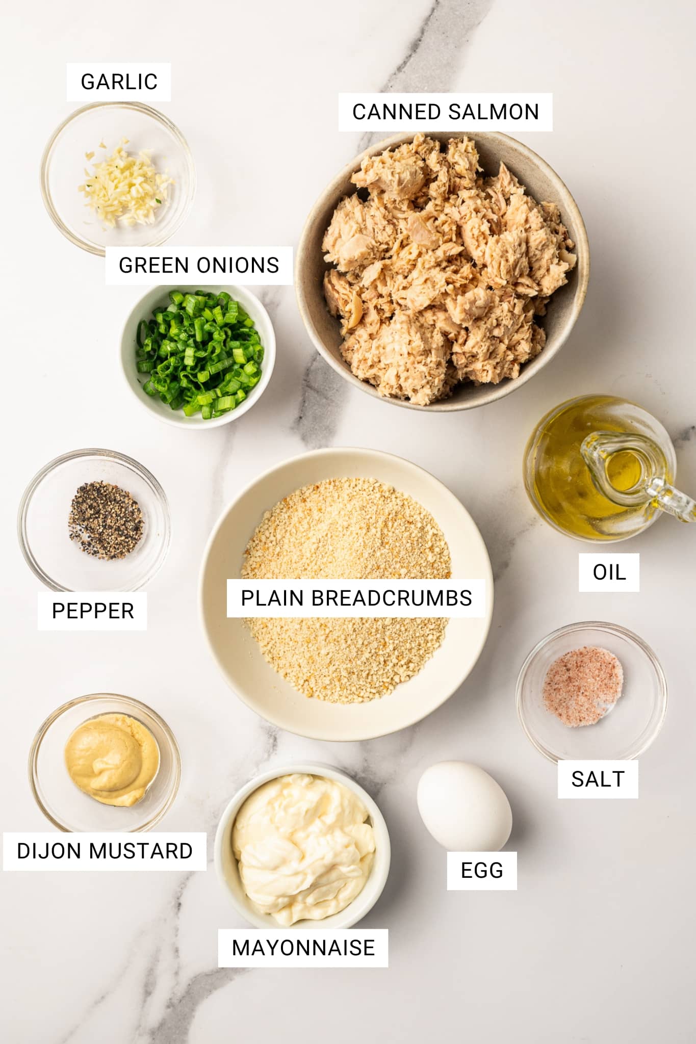 Overhead image of salmon cake ingredients arranged on a white marble surface with text labels. The labeled ingredients include garlic, canned salmon, oil, green onions, pepper, plain breadcrumbs, salt, Dijon mustard, mayonnaise, and egg. The ingredients are shown in small glass bowls, a beige bowl, a cream bowl, and a glass oil cruet.