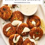 Pinterest pin showing crispy, golden-brown salmon cakes on a cream-colored oval platter with creamy white sauce drizzled over the tops, chopped green chives, a bowl of creamy sauce, lemon wedges, and an olive-green napkin. A white rectangular text overlay at the top reads "SALMON CAKES" in large black letters, "(WITH CANNED SALMON)" below it, a thin yellow line, and "THEFORKEDSPOON.COM."