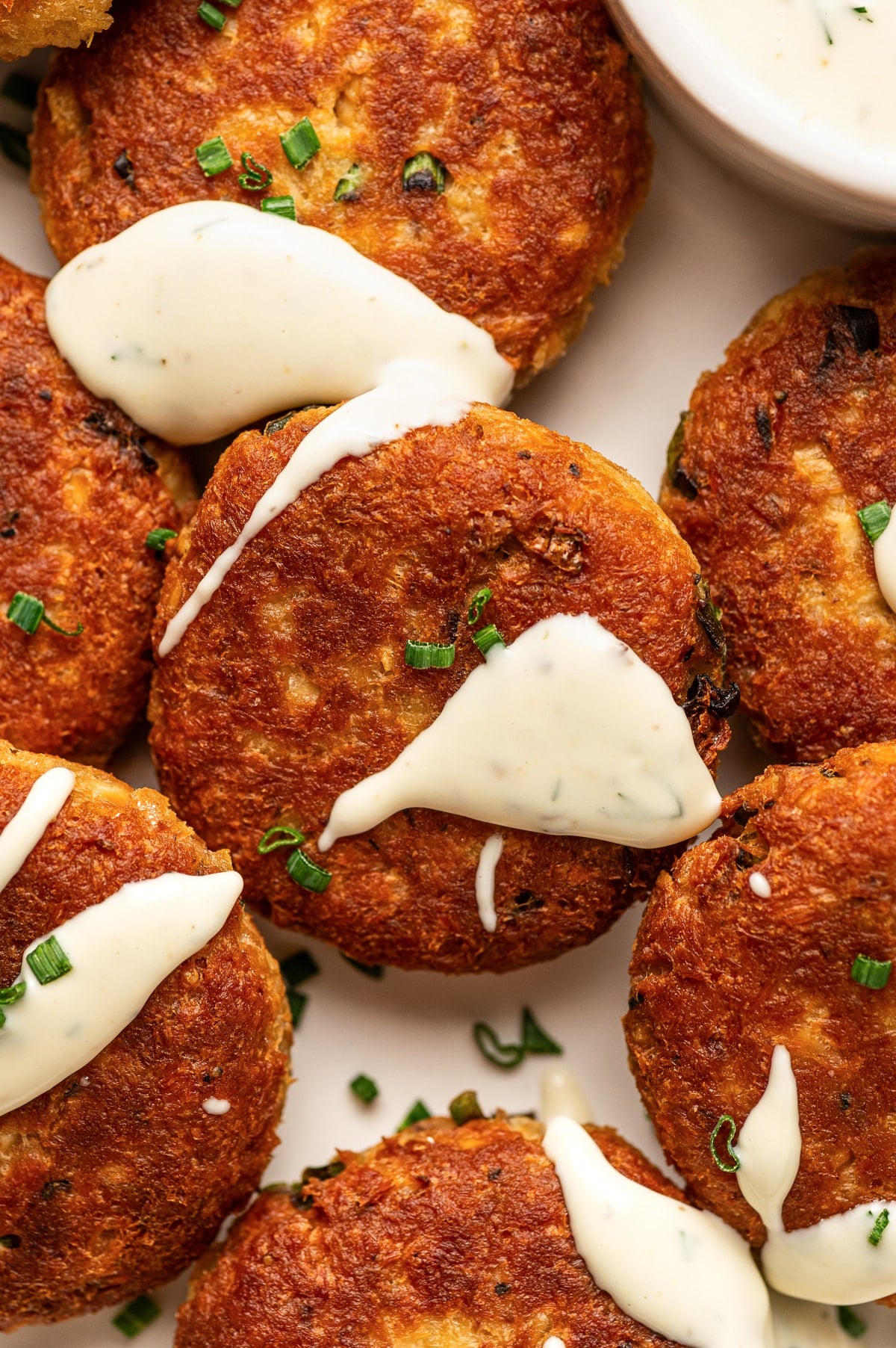 Close-up overhead view of crispy golden salmon cakes drizzled with creamy white sauce and sprinkled with chopped green chives. A small bowl of sauce is partially visible in the background.