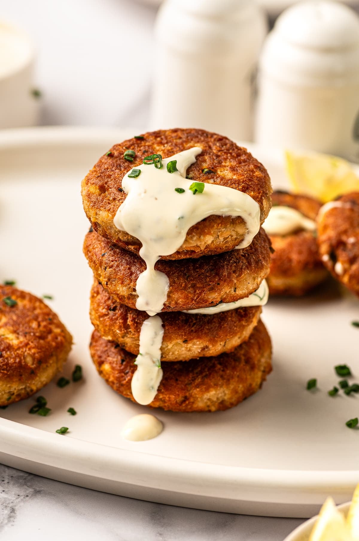 Stack of crispy golden salmon cakes on a cream platter with creamy white sauce dripping down the sides, topped with chopped green chives, with more salmon cakes, lemon wedges, and small white condiment jars softly blurred in the background.