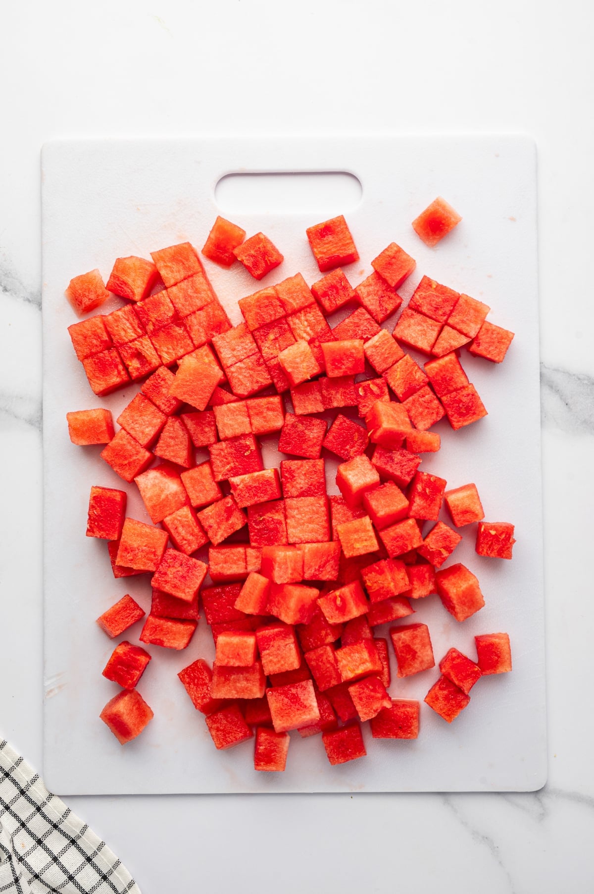 Cubed red watermelon piled on a white cutting board on a white marble countertop, with a black and white checkered kitchen towel in the lower left corner.