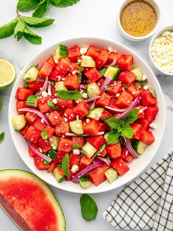 Finished watermelon cucumber feta salad in a large white bowl with cubed red watermelon, chopped cucumber, sliced red onion, crumbled feta cheese, fresh mint, basil, and black pepper, served with lime halves, a bowl of dressing, extra feta, fresh mint, a watermelon wedge, and a black and white checkered towel on a white marble countertop.