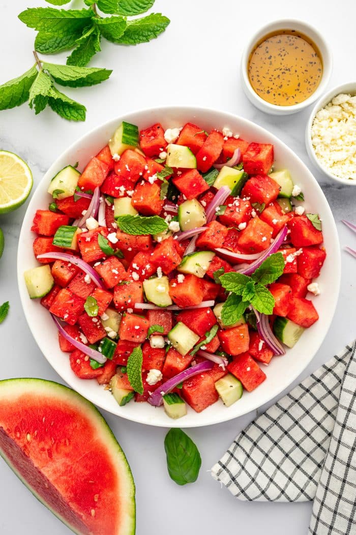 Finished watermelon cucumber feta salad in a large white bowl with cubed red watermelon, chopped cucumber, sliced red onion, crumbled feta cheese, fresh mint, basil, and black pepper, served with lime halves, a bowl of dressing, extra feta, fresh mint, a watermelon wedge, and a black and white checkered towel on a white marble countertop.