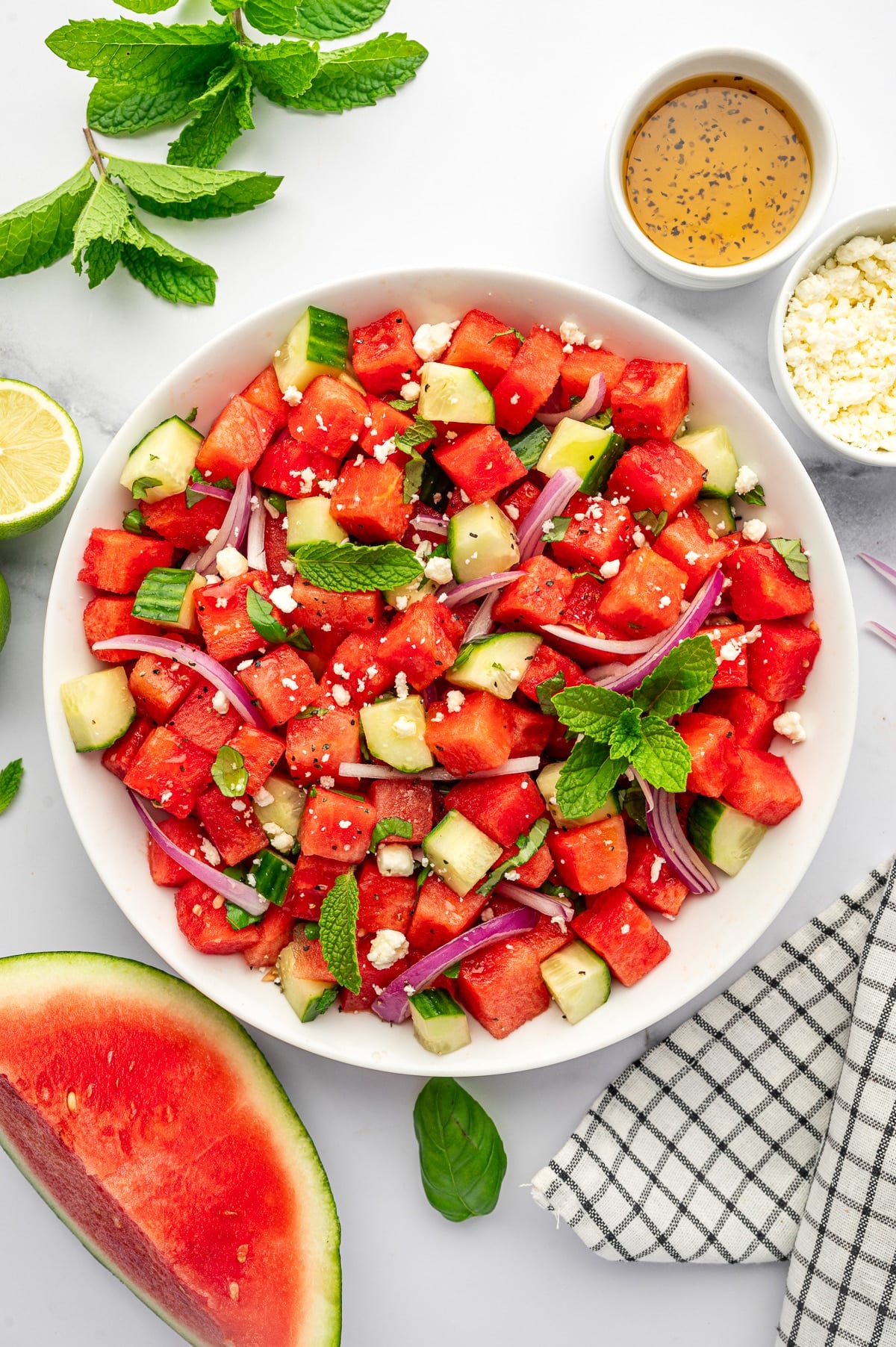 Finished watermelon cucumber feta salad in a large white bowl with cubed red watermelon, chopped cucumber, sliced red onion, crumbled feta cheese, fresh mint, basil, and black pepper, served with lime halves, a bowl of dressing, extra feta, fresh mint, a watermelon wedge, and a black and white checkered towel on a white marble countertop.
