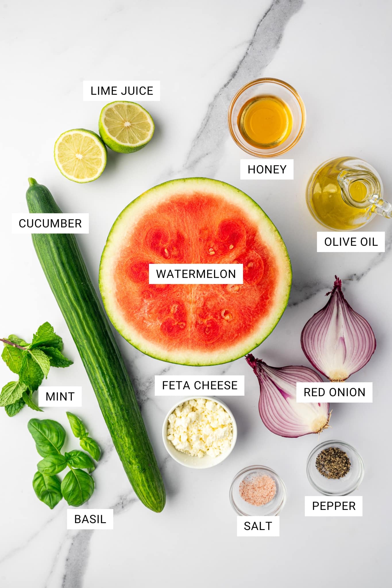 Watermelon salad ingredients arranged on a white marble countertop with text labels, including lime juice, honey, olive oil, watermelon, cucumber, red onion, feta cheese, mint, basil, salt, and pepper.