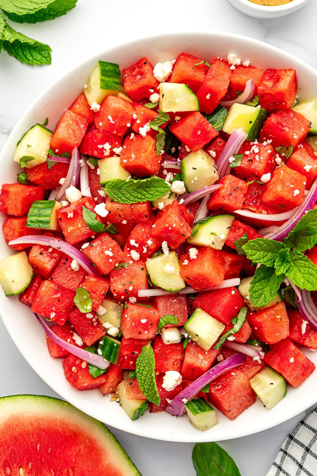 Close-up of watermelon salad in a white bowl with cubed red watermelon, chopped cucumber, thin red onion slices, crumbled feta cheese, fresh mint leaves, basil, and black pepper, with a watermelon wedge and black and white checkered towel nearby.