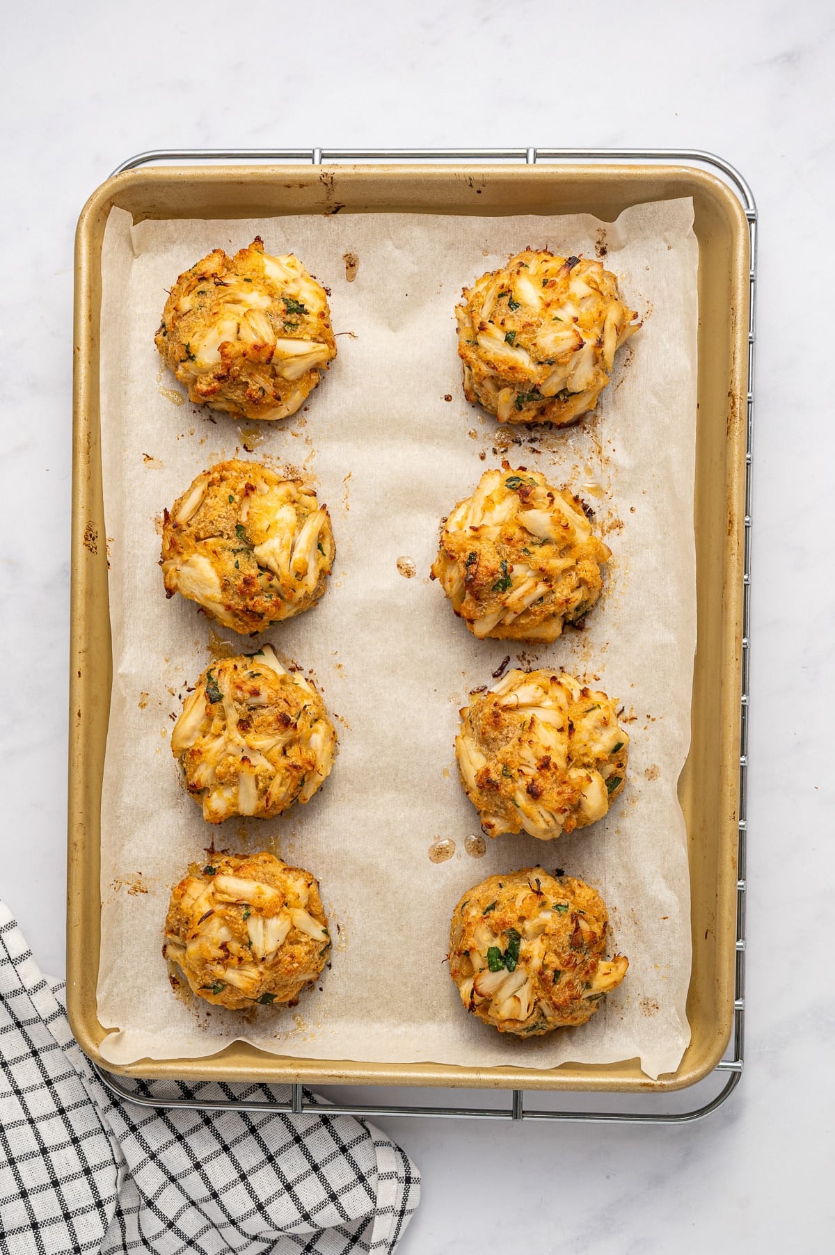 Eight golden, baked Maryland-style crab cakes on a parchment-lined gold baking sheet set over a cooling rack, with browned edges, visible lump crab meat, parsley, and a black-and-white checkered towel nearby.