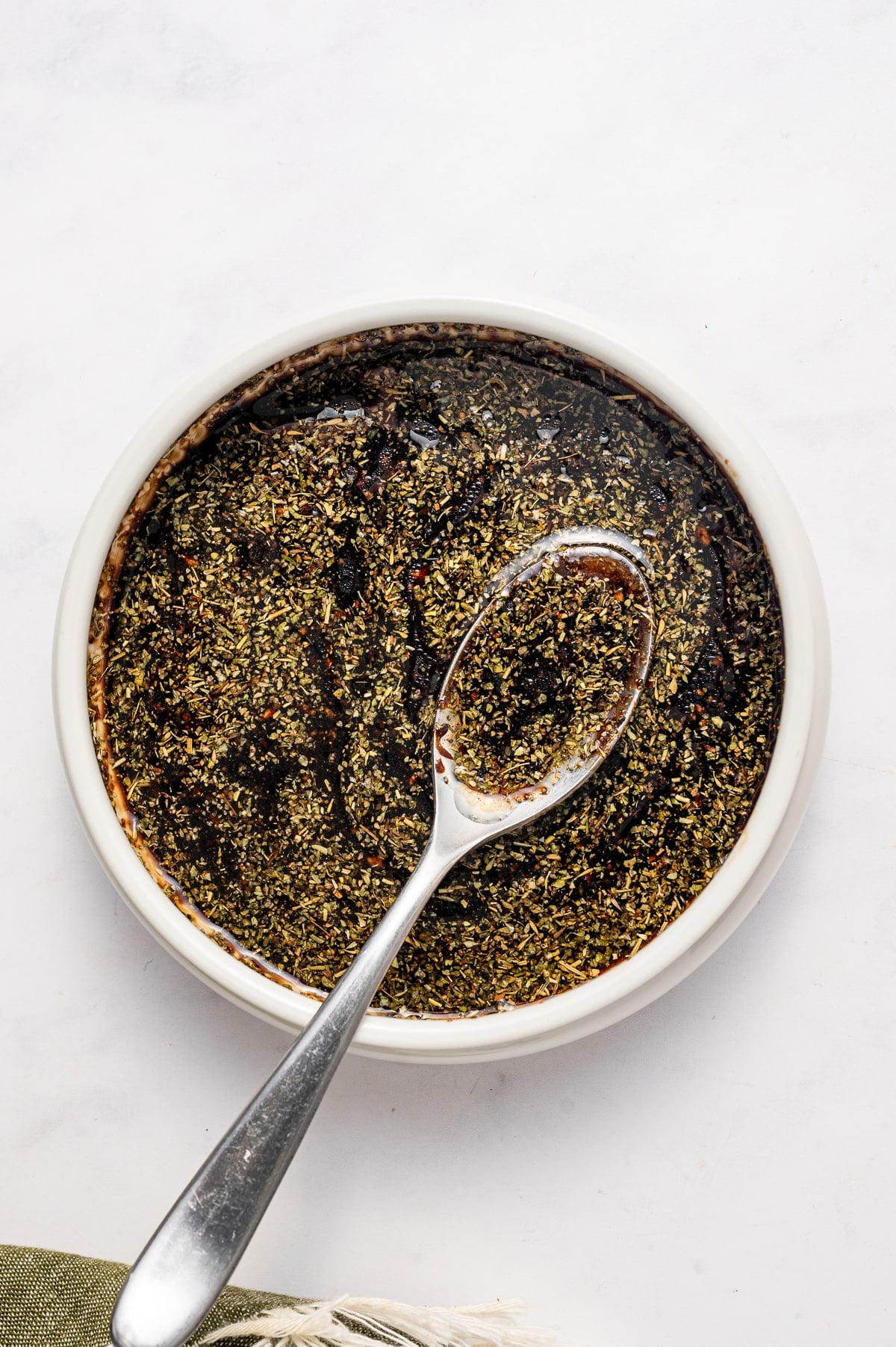 Dark balsamic chicken marinade with visible Italian seasoning and black pepper in a small round white bowl, with a silver spoon resting in the marinade on a white marble surface.