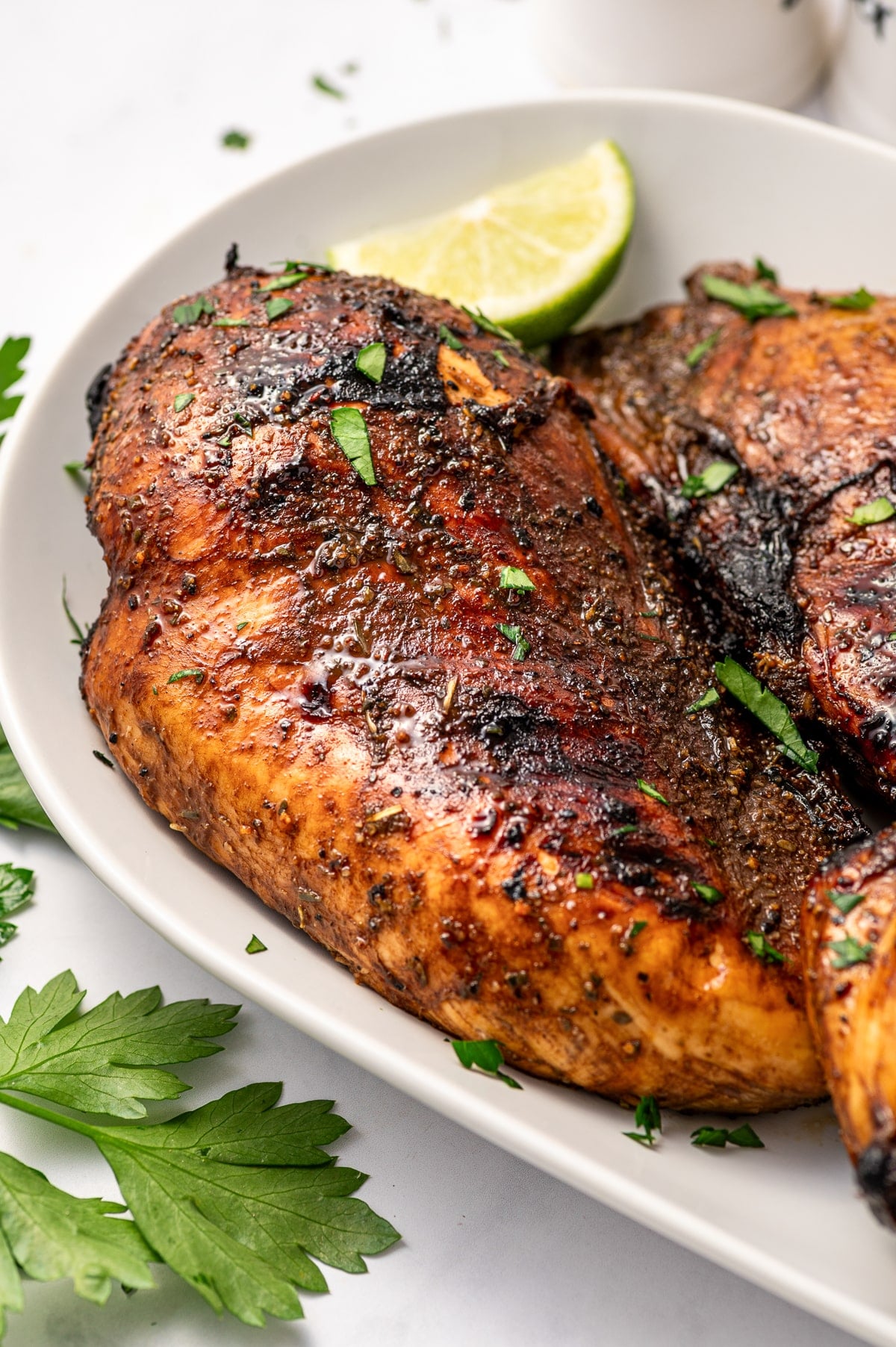 Close-up of two grilled chicken breasts with dark balsamic glaze, charred grill marks, chopped parsley, and a lime wedge on a white oval serving platter.