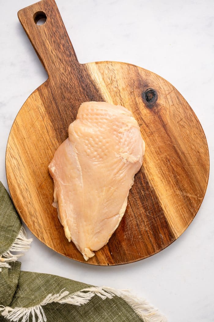 Two raw boneless, skinless chicken breasts on a round wooden cutting board with a long handle, set on a white marble surface beside an olive green fringed kitchen towel.