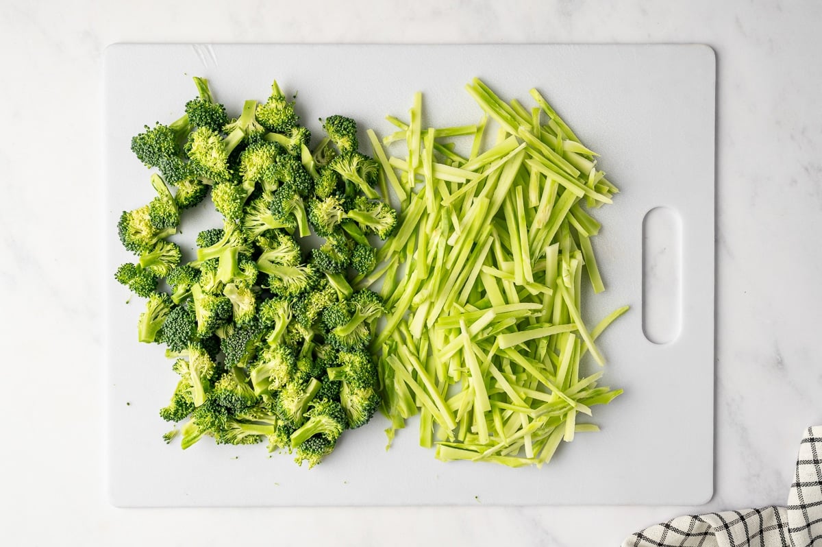 Chopped broccoli florets and thinly sliced broccoli stems arranged side by side on a white cutting board on a white marble surface, with a black-and-white kitchen towel in the corner.