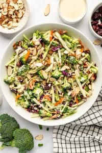 Finished broccoli slaw in a large white oval serving bowl with broccoli florets, thin broccoli stems, red cabbage, carrots, dried cranberries, sliced almonds, and creamy dressing, surrounded by small bowls of almonds, dressing, and cranberries.