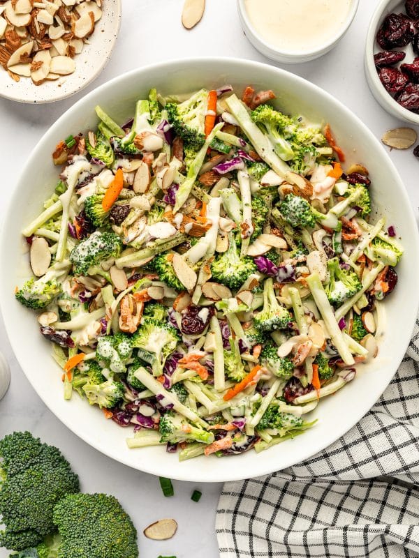 Finished broccoli slaw in a large white oval serving bowl with broccoli florets, thin broccoli stems, red cabbage, carrots, dried cranberries, sliced almonds, and creamy dressing, surrounded by small bowls of almonds, dressing, and cranberries.