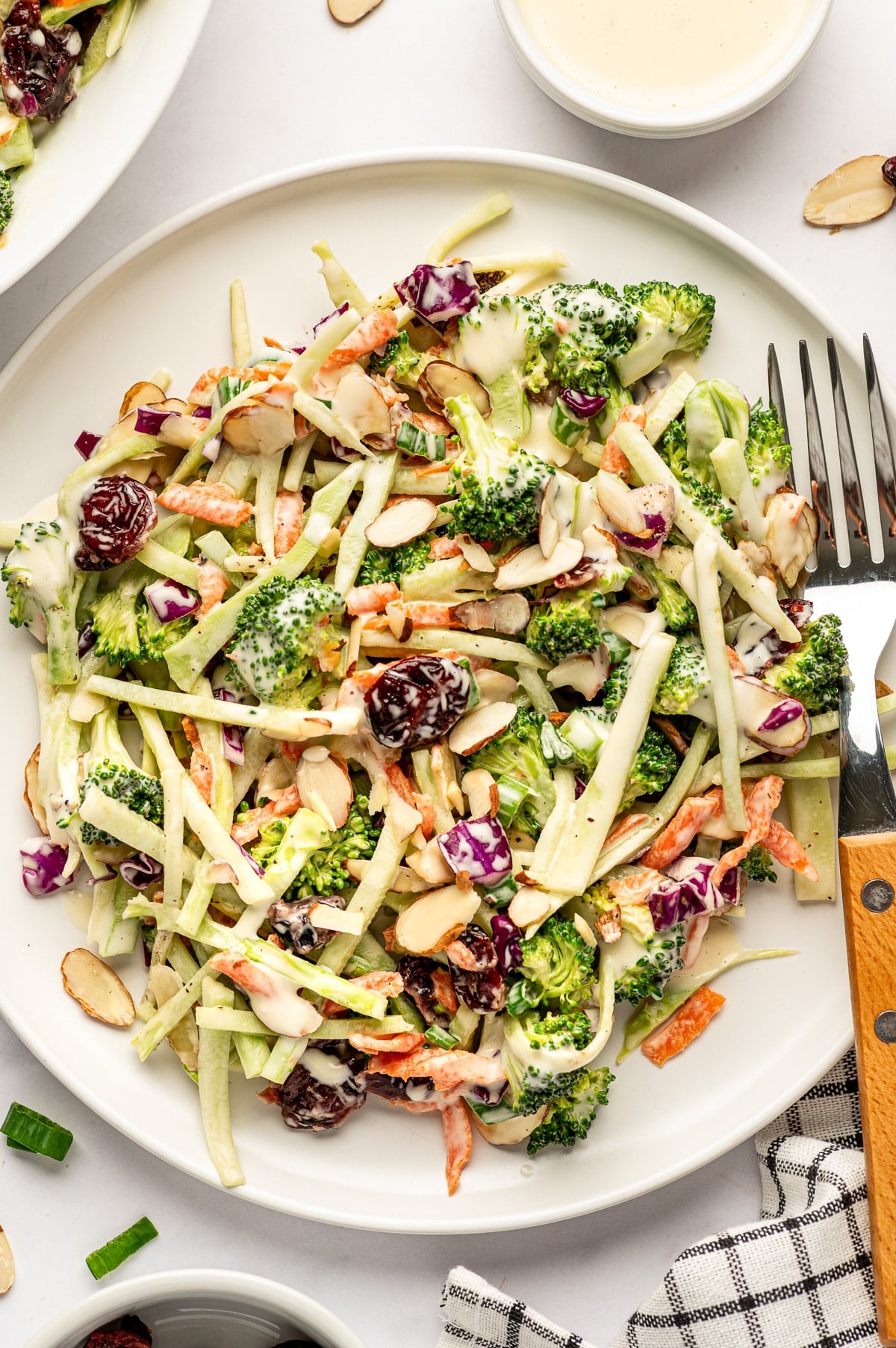 A serving of broccoli slaw on a small white plate with a large fork, featuring broccoli florets, thin broccoli stems, carrots, red cabbage, cranberries, sliced almonds, and creamy dressing.