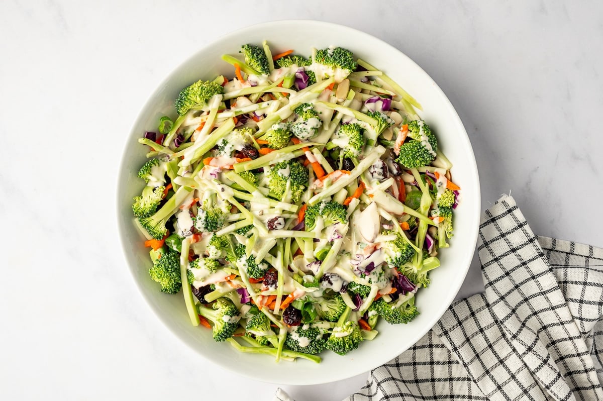 Fresh broccoli slaw in a large white oval bowl with broccoli florets, broccoli stems, red cabbage, carrots, cranberries, and almonds drizzled with creamy dressing.