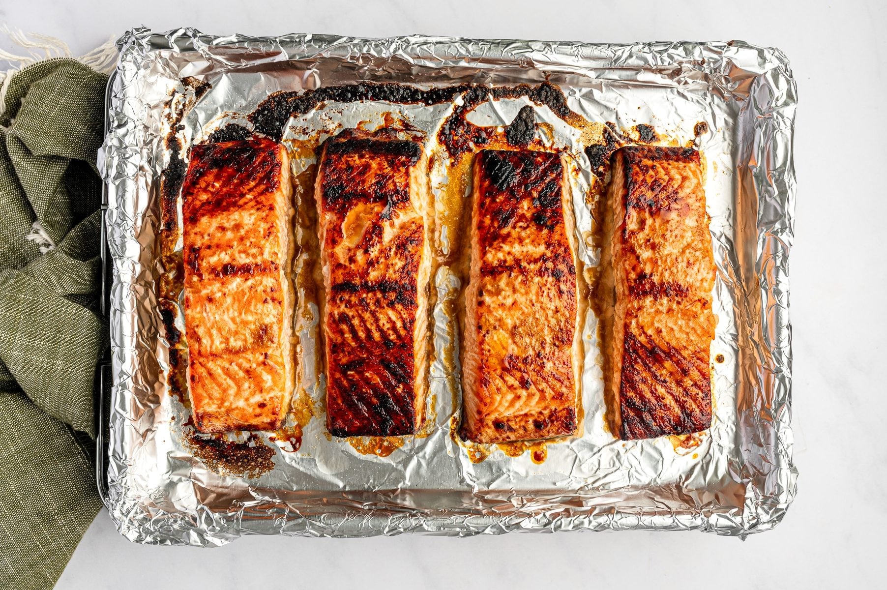 Four broiled teriyaki salmon fillets on a foil-lined baking sheet with caramelized browned edges, dark charred glaze spots, and a green cloth napkin at the bottom.