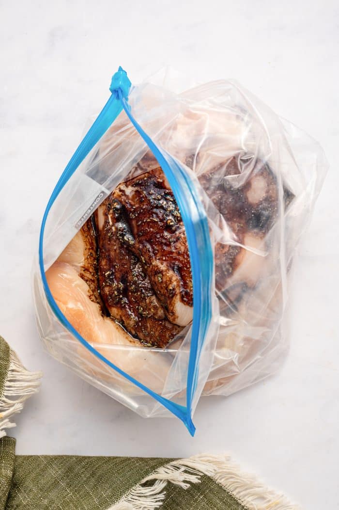 Raw chicken breasts coated in dark balsamic marinade inside an open clear zip-top bag with bright blue zipper edges, set on a white marble surface beside an olive green fringed towel.