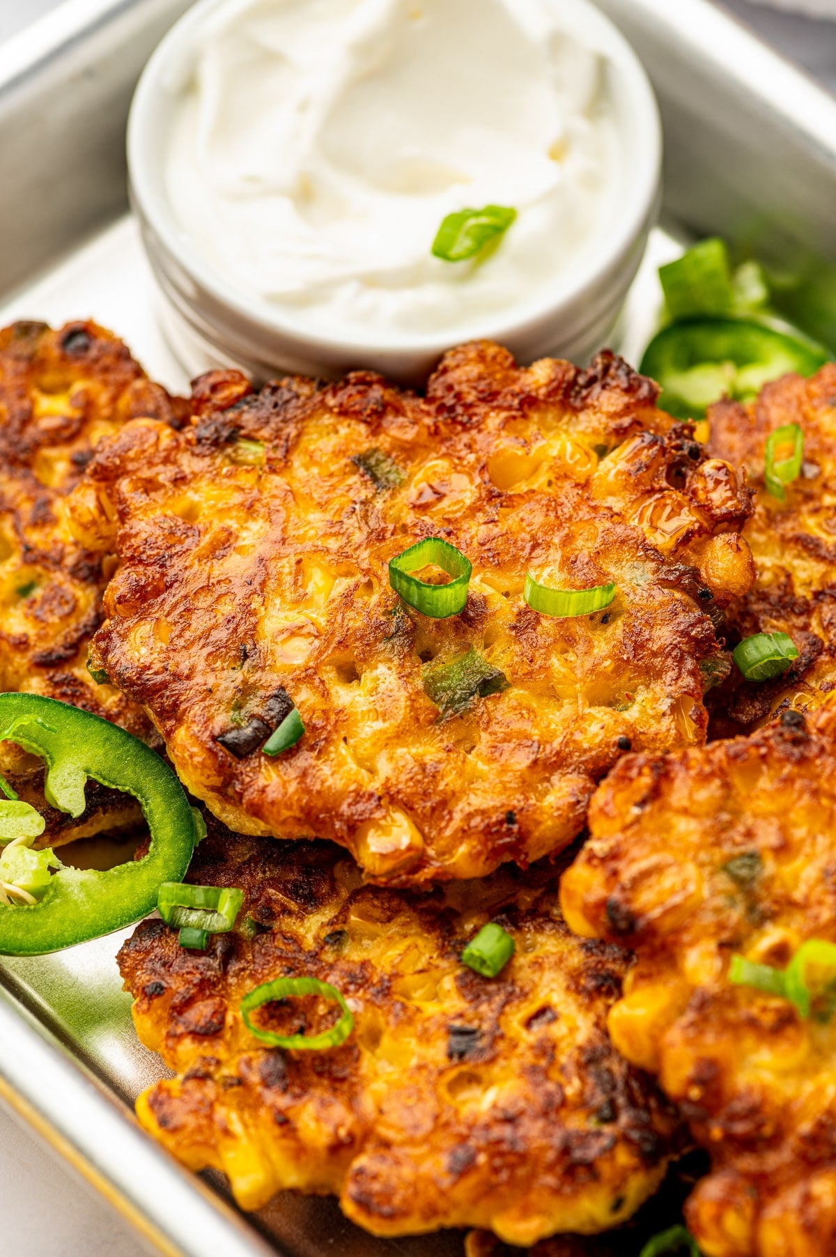 Close-up of crispy corn fritters with browned edges, visible corn kernels, green onions, jalapeños, and a small bowl of sour cream in the background.