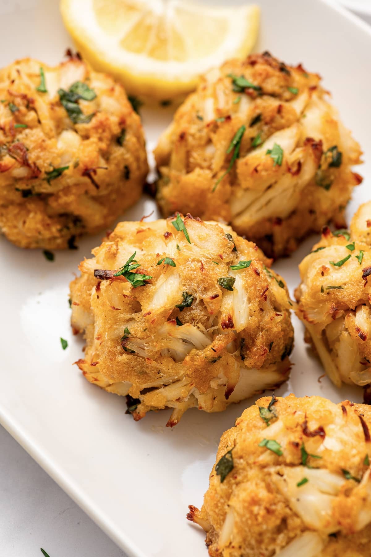 Close-up of golden Maryland-style crab cakes with large visible pieces of lump crab meat, chopped parsley, and browned edges served on a white plate with a lemon wedge in the background.