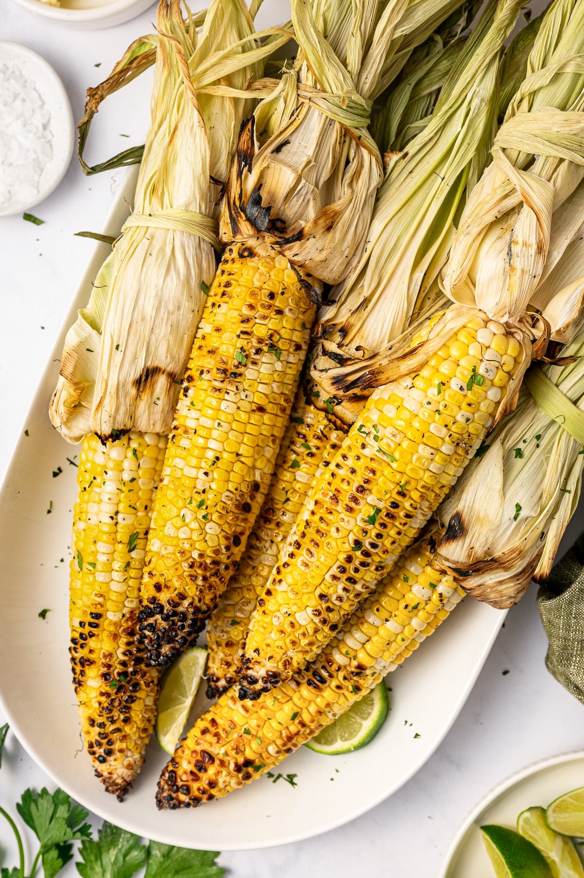 Close-up of grilled corn on the cob with husks tied back on a white oval platter, garnished with chopped green herbs and served with lime wedges.