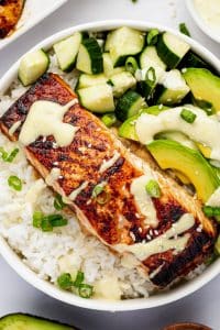 Close-up of a white bowl filled with teriyaki salmon over white rice, diced cucumber, sliced avocado, green onion, sesame seeds, and creamy wasabi mayo.