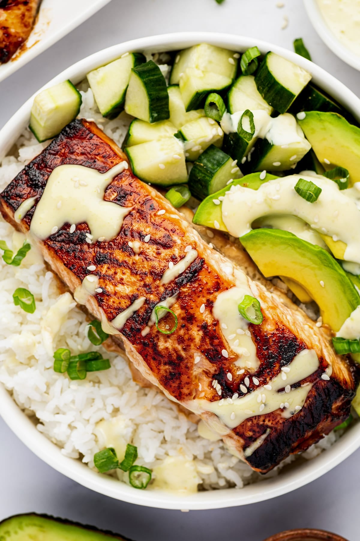 Close-up of a white bowl filled with teriyaki salmon over white rice, diced cucumber, sliced avocado, green onion, sesame seeds, and creamy wasabi mayo.