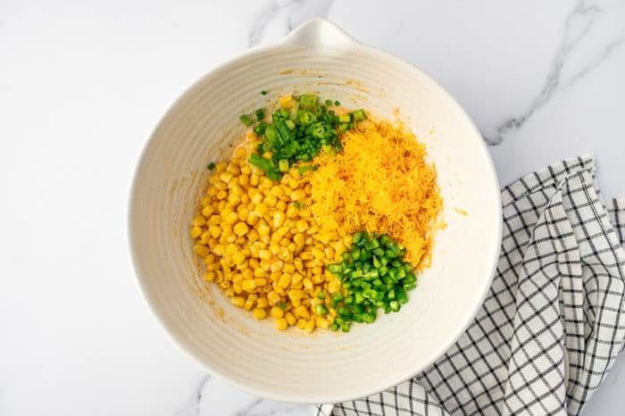 Corn kernels, shredded cheddar cheese, chopped green onions, and diced jalapeños were added to the corn fritter batter in a cream-colored mixing bowl.