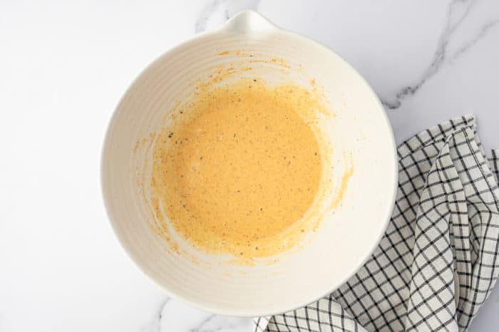 Smooth yellow corn fritter batter mixed in a cream-colored bowl with visible flecks of paprika and black pepper on a white marble surface.