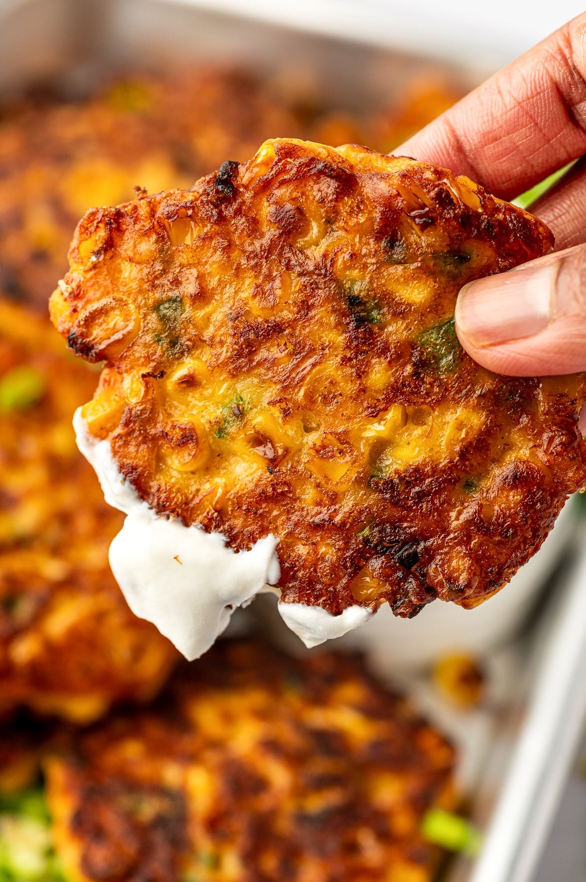 A hand holding a crispy corn fritter dipped in sour cream, showing golden browned edges, corn kernels, and green onion pieces.