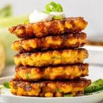 Pinterest pin image showing a stack of golden corn fritters on a white plate with sour cream, green onions, and jalapeรฑos, plus text overlay reading "CORN FRITTERS" and TheForkedSpoon.com.