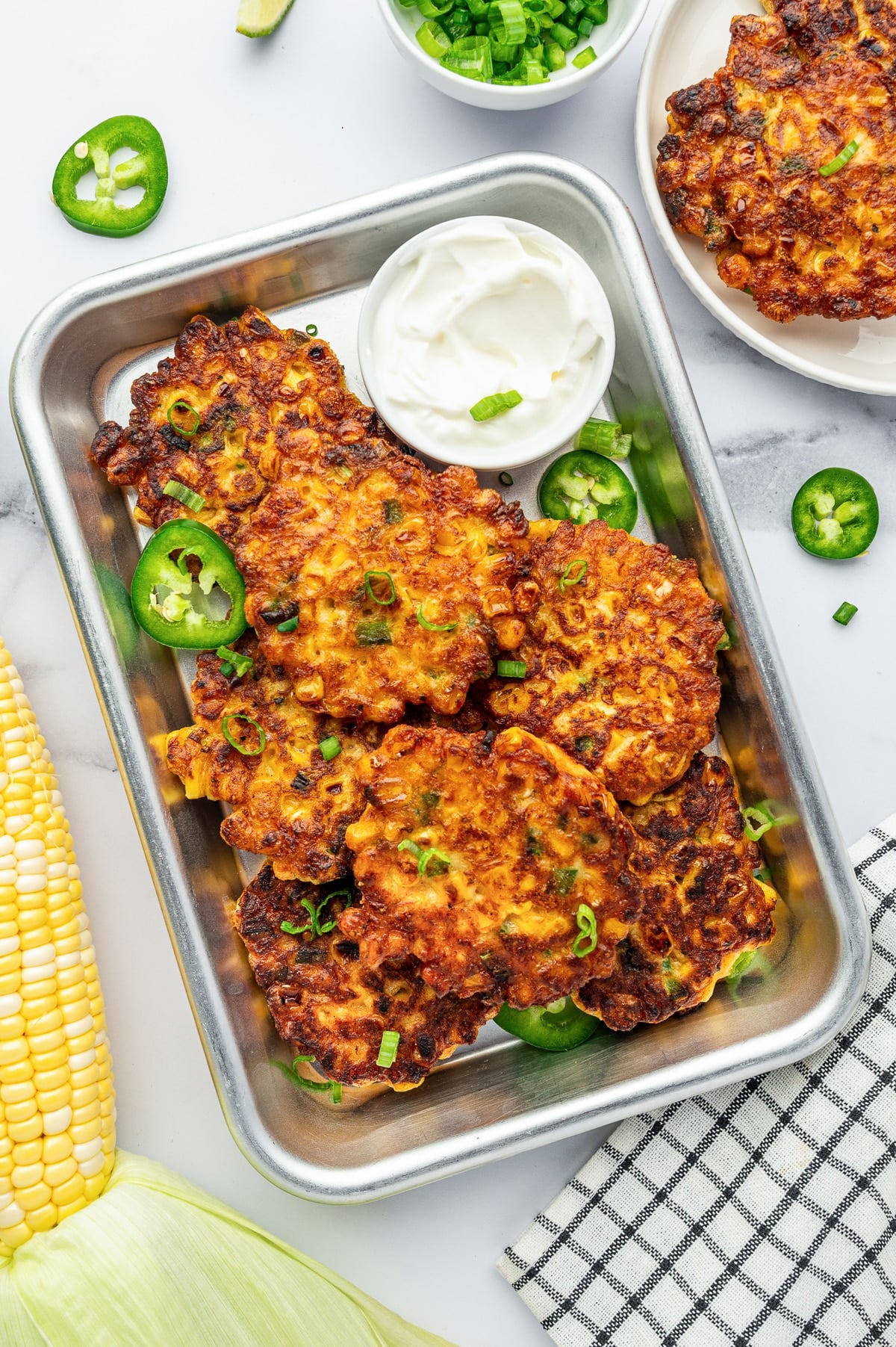 Crispy golden corn fritters served in a small metal tray with sour cream, sliced jalapeños, green onions, and fresh corn on the cob nearby.