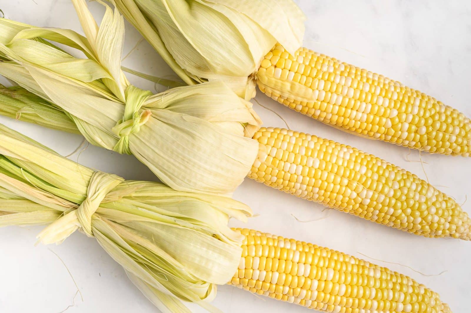 Three ears of yellow corn with the husks peeled back and tied near the base, arranged on a white marble surface.