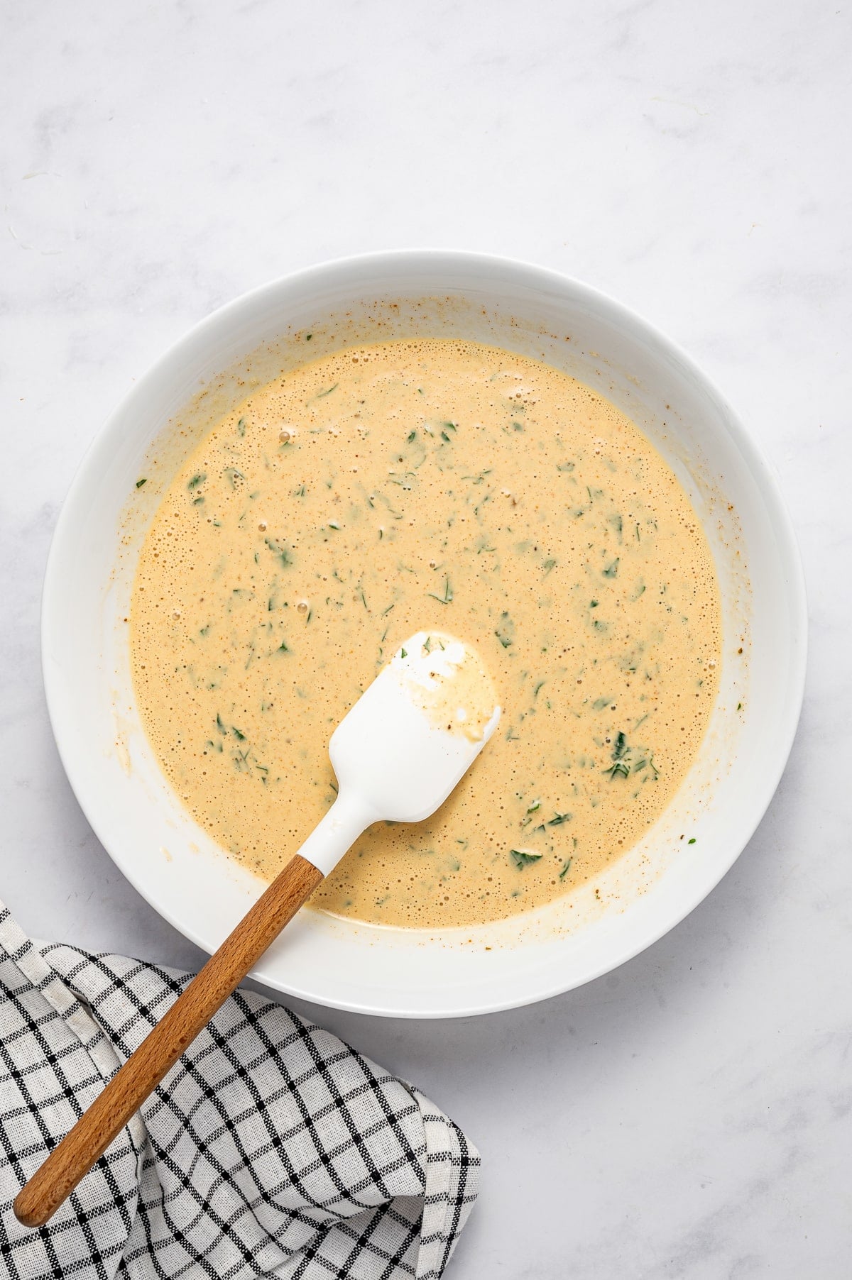 Creamy orange crab cake binder mixture with chopped parsley in a large white bowl, stirred with a white spatula with a wooden handle on a white marble surface beside a black-and-white checkered towel.