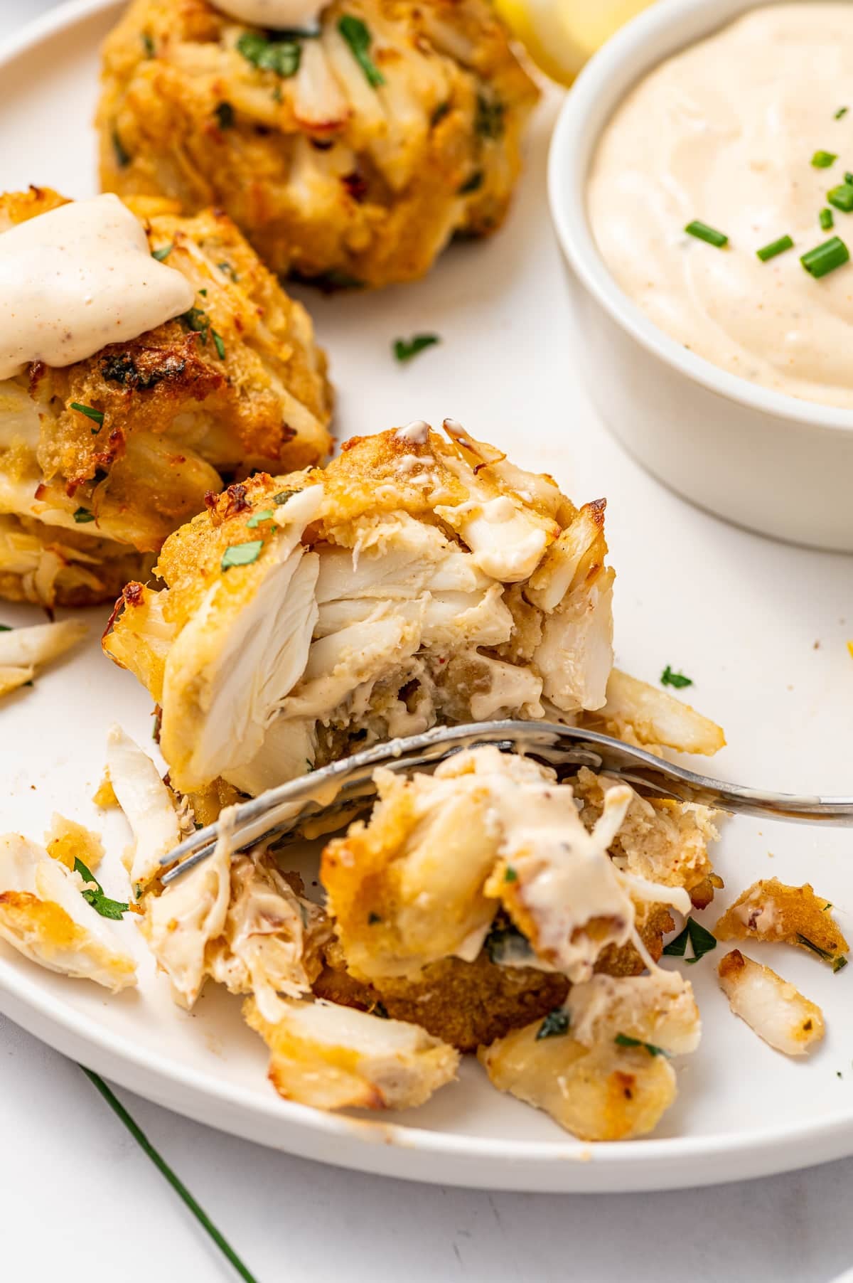 Close-up of a fork cutting into a golden crab cake, showing the moist interior with large pieces of lump crab meat, creamy sauce, chopped parsley, lemon wedges, and a white ramekin of dipping sauce in the background.
