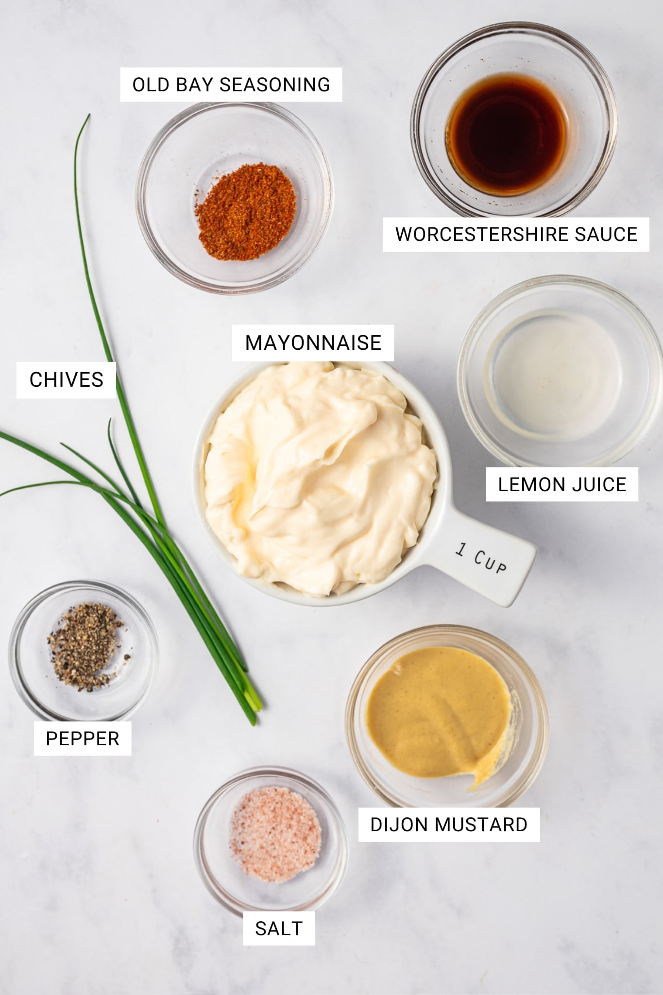 Labeled overhead photo of the ingredients for crab cake sauce showing Old Bay seasoning, Worcestershire sauce, mayonnaise in a white measuring cup, lemon juice, Dijon mustard, salt, pepper, and fresh chives on a light surface.