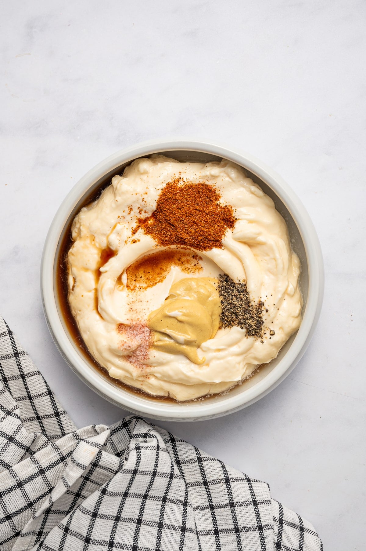 Overhead view of crab cake sauce ingredients in a white bowl, including mayonnaise, Dijon mustard, Old Bay seasoning, Worcestershire sauce, salt, and black pepper, with a black-and-white checkered towel on a light surface.