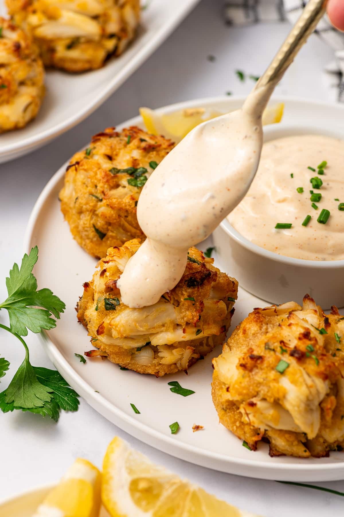 Creamy crab cake sauce being spooned over a golden crab cake on a white plate, with more crab cakes, a white ramekin of sauce, lemon wedges, parsley, and chopped chives around it.
