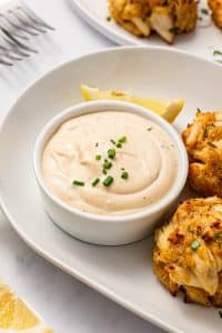Creamy crab cake sauce in a white ramekin on a white oval plate, served with golden Maryland-style crab cakes, lemon wedges, chopped chives, and forks in the background.