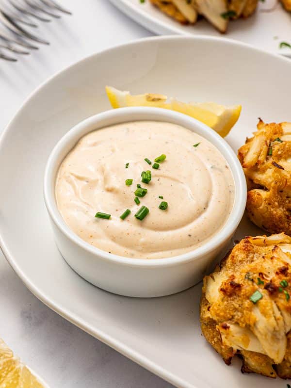 Creamy crab cake sauce in a white ramekin on a white oval plate, served with golden Maryland-style crab cakes, lemon wedges, chopped chives, and forks in the background.