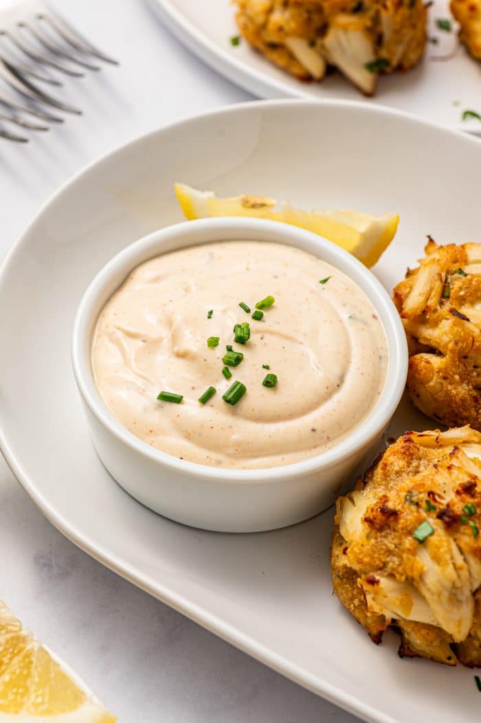 Creamy crab cake sauce in a white ramekin on a white oval plate, served with golden Maryland-style crab cakes, lemon wedges, chopped chives, and forks in the background.