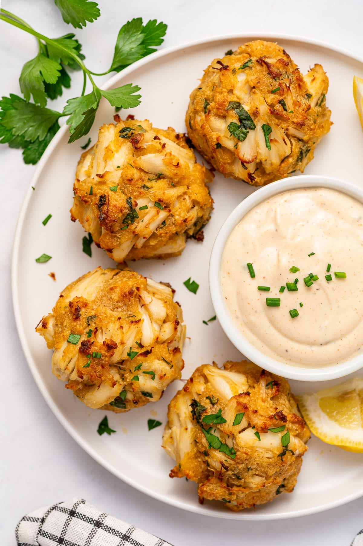 Close-up overhead image of four golden crab cakes on a white plate with visible lump crab meat, chopped parsley, lemon wedges, and a white ramekin of creamy dipping sauce garnished with chives.