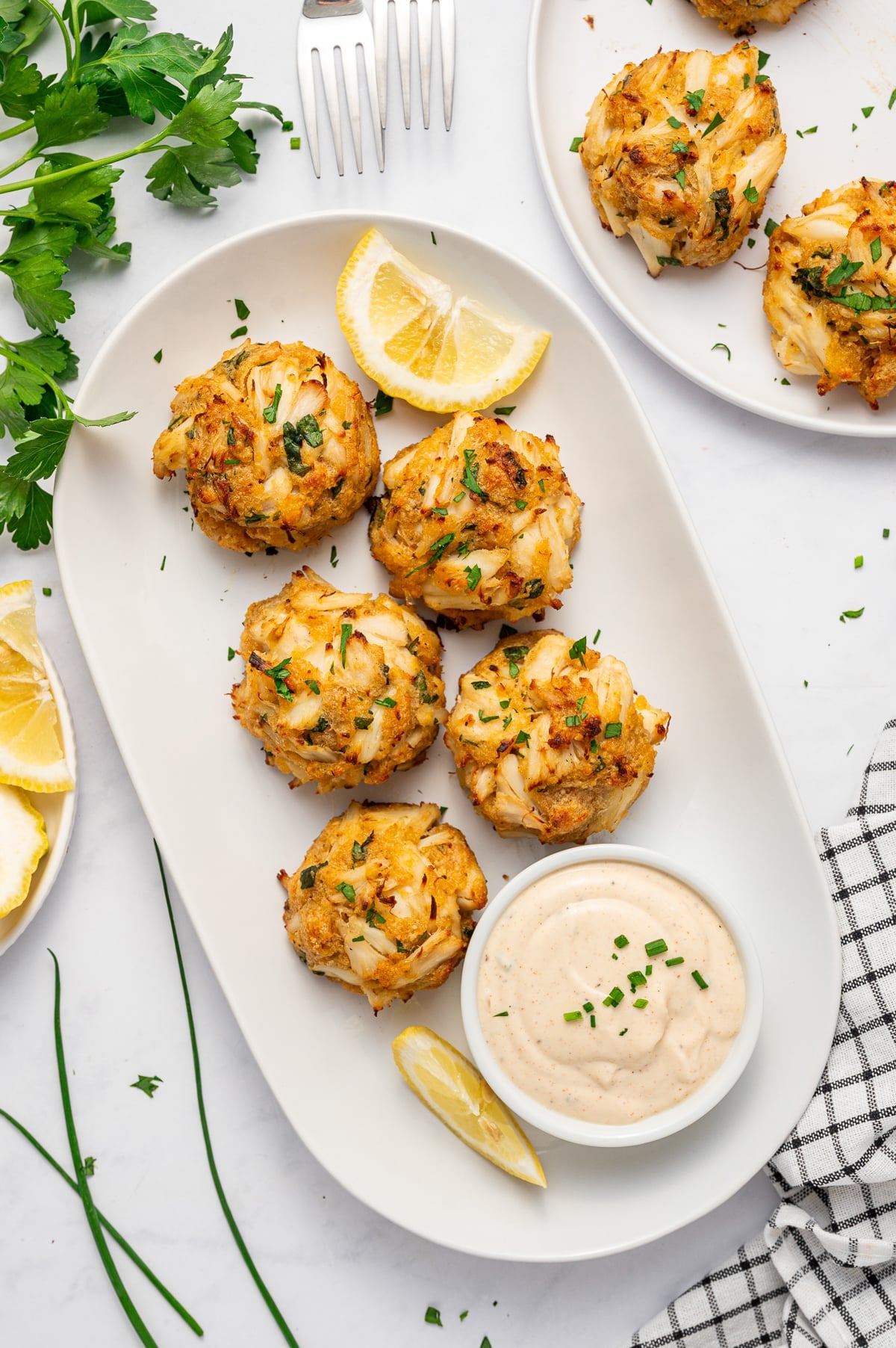 Six golden crab cakes with visible pieces of lump crab meat served on a white oval platter with lemon wedges, chopped parsley, and a small white bowl of creamy sauce garnished with chives, surrounded by fresh parsley, lemon wedges, a fork, and a black and white checkered towel.